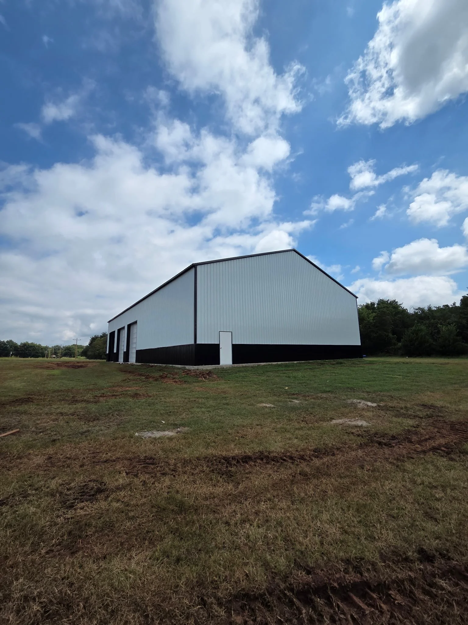 Large white metal building with black trim sitting on grassy land under a cloudy blue sky.