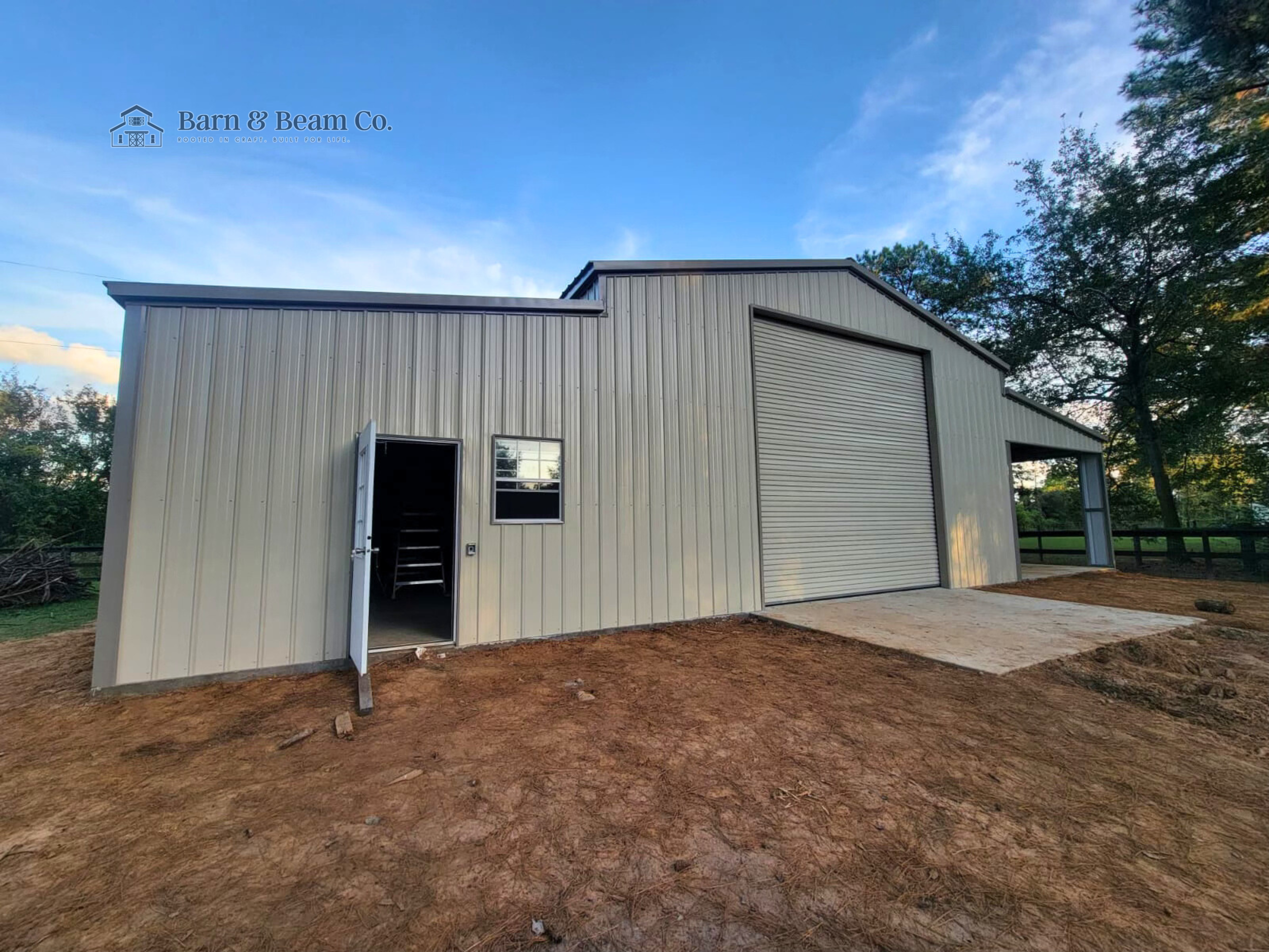 Metal building with a roll-up door, a small side door, and a window, situated on a patch of dirt, with trees and blue sky in the background.