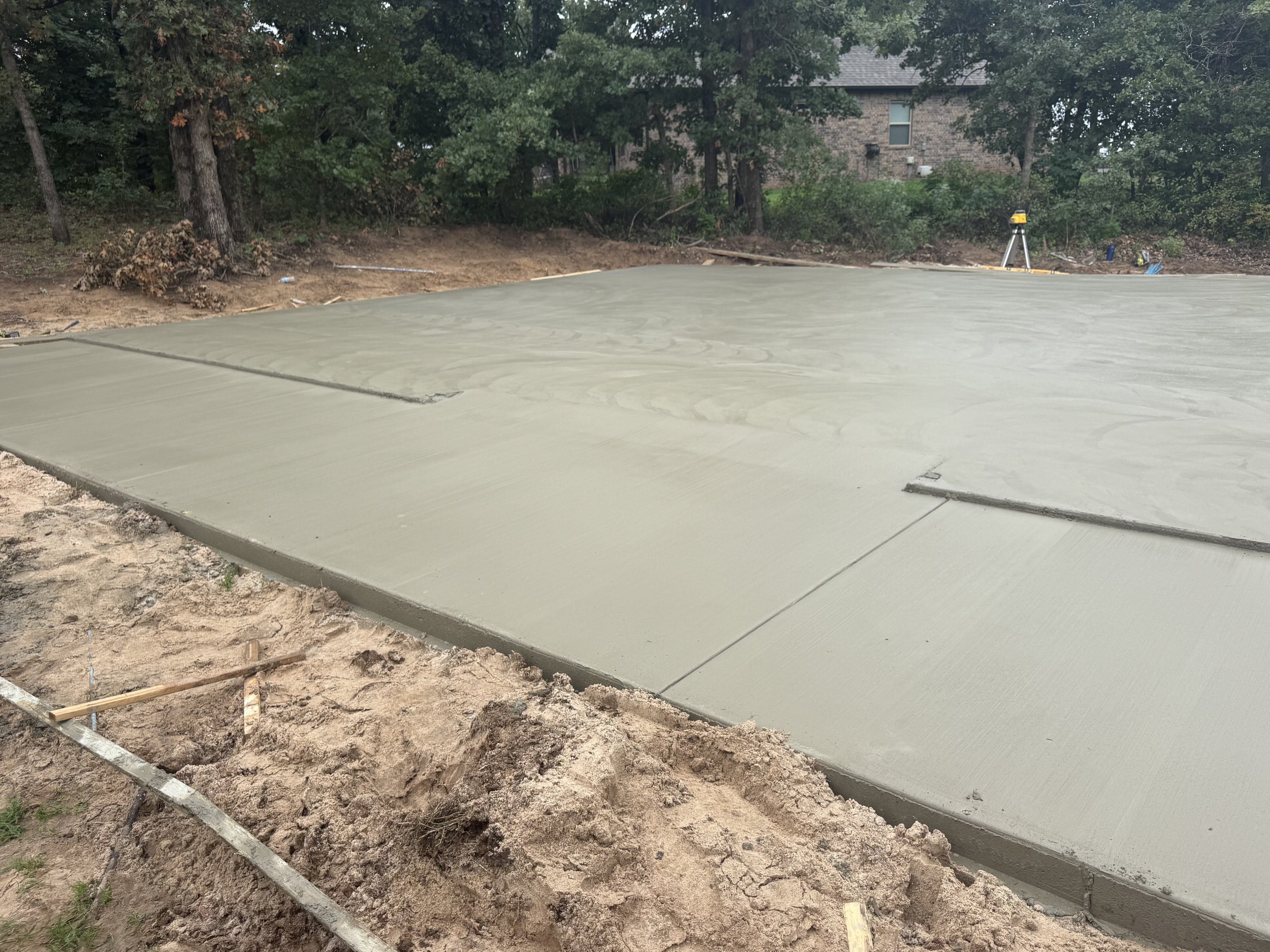 Freshly poured concrete slab on a construction site surrounded by dirt and trees, with a house and construction equipment in the background.