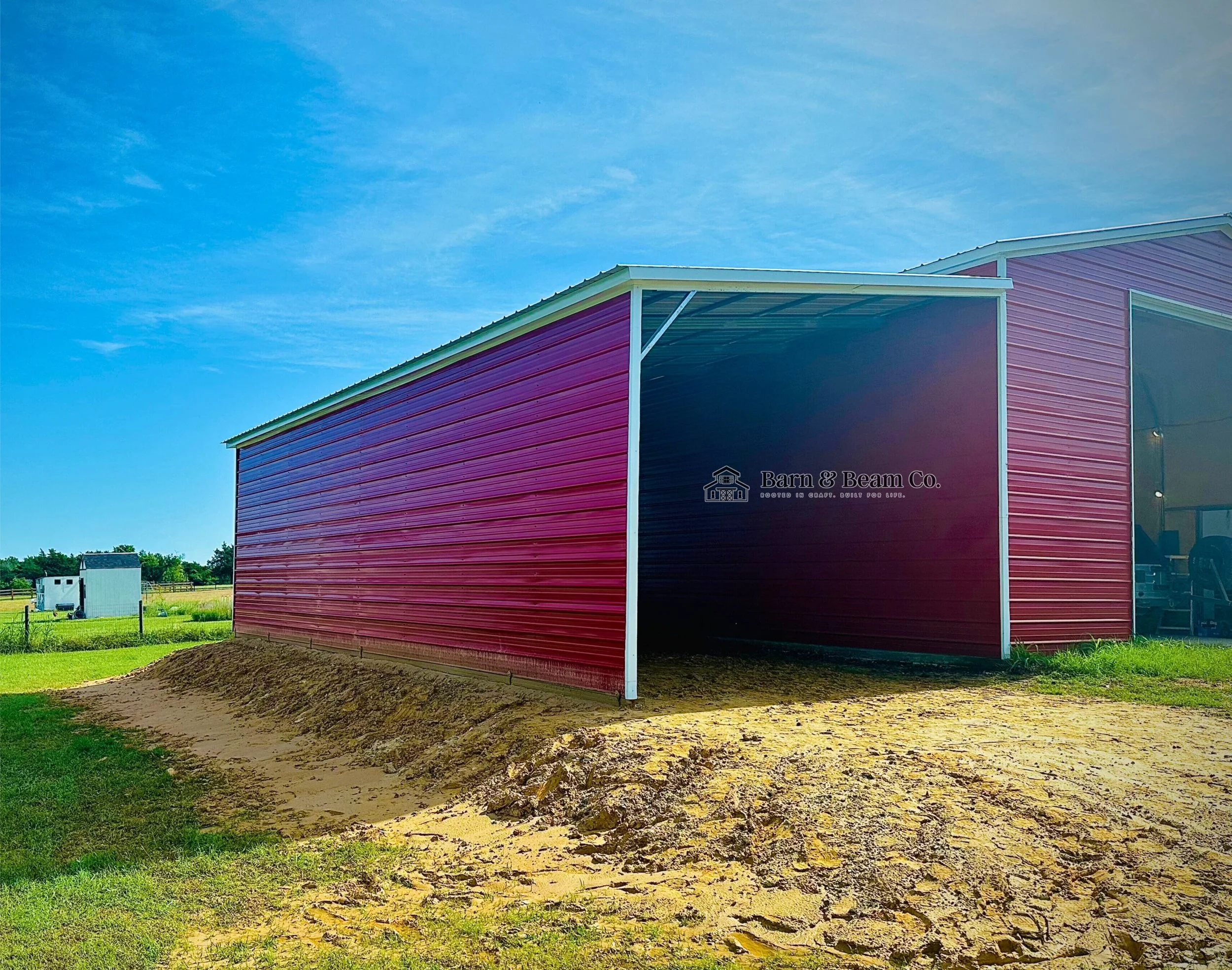 Red metal barn with open entrance on a farm with green grass, a fenced area, and small white buildings in background under a blue sky.