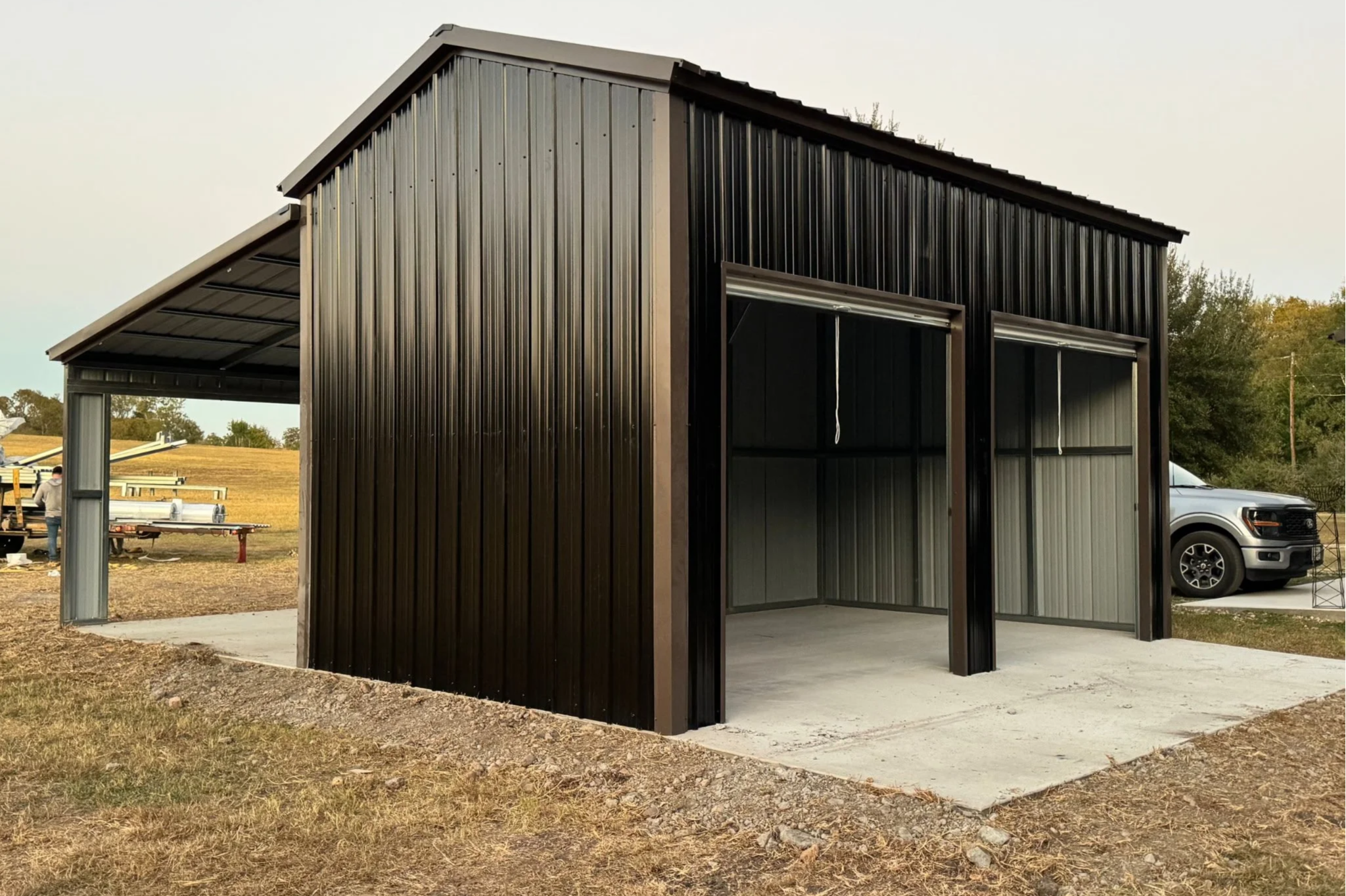 Black metal storage shed with two open garage doors, concrete floor, and an adjacent covered area