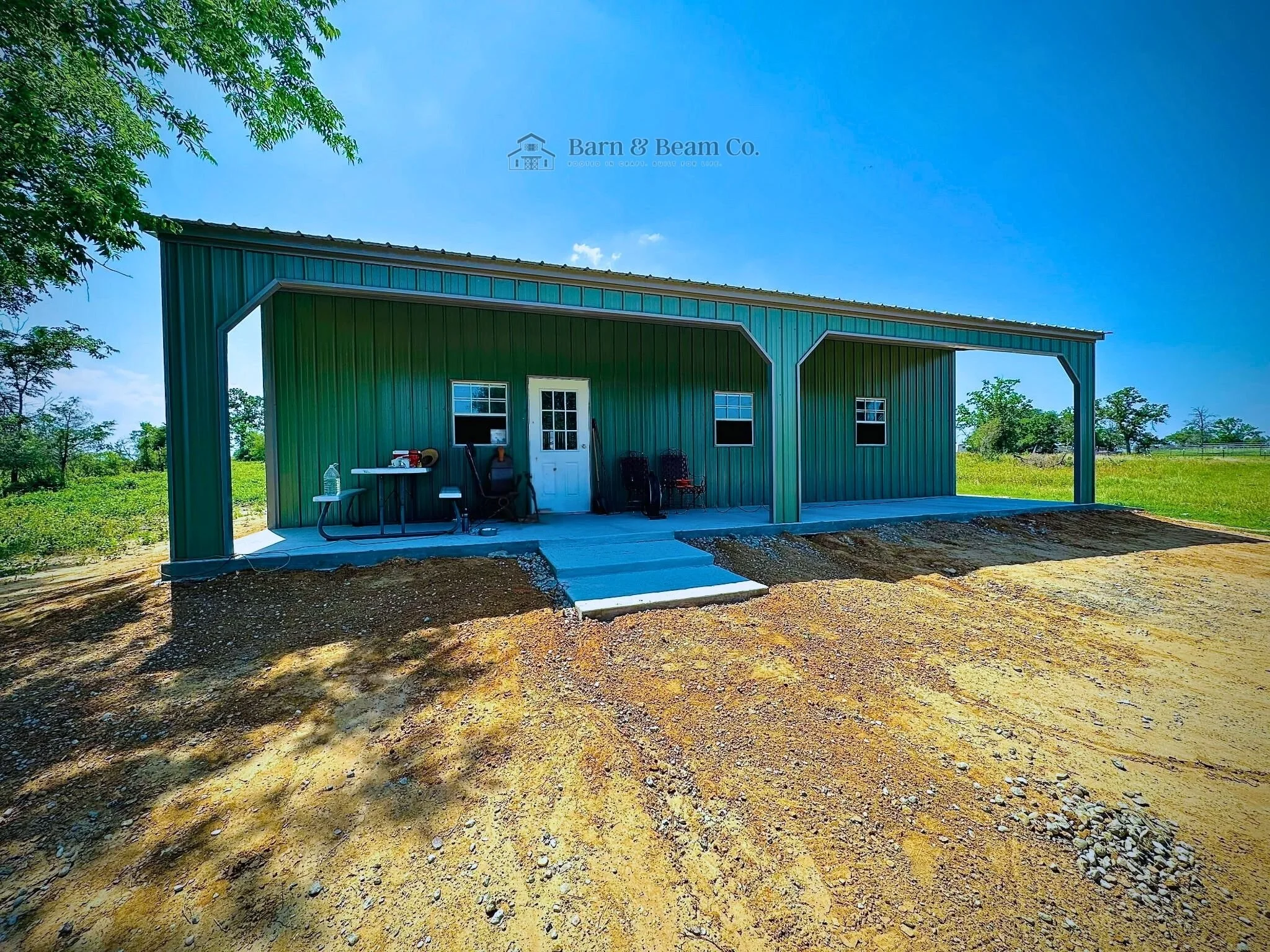A green metal building with an open carport, located in a rural area with trees and open land in the background, under a clear blue sky.