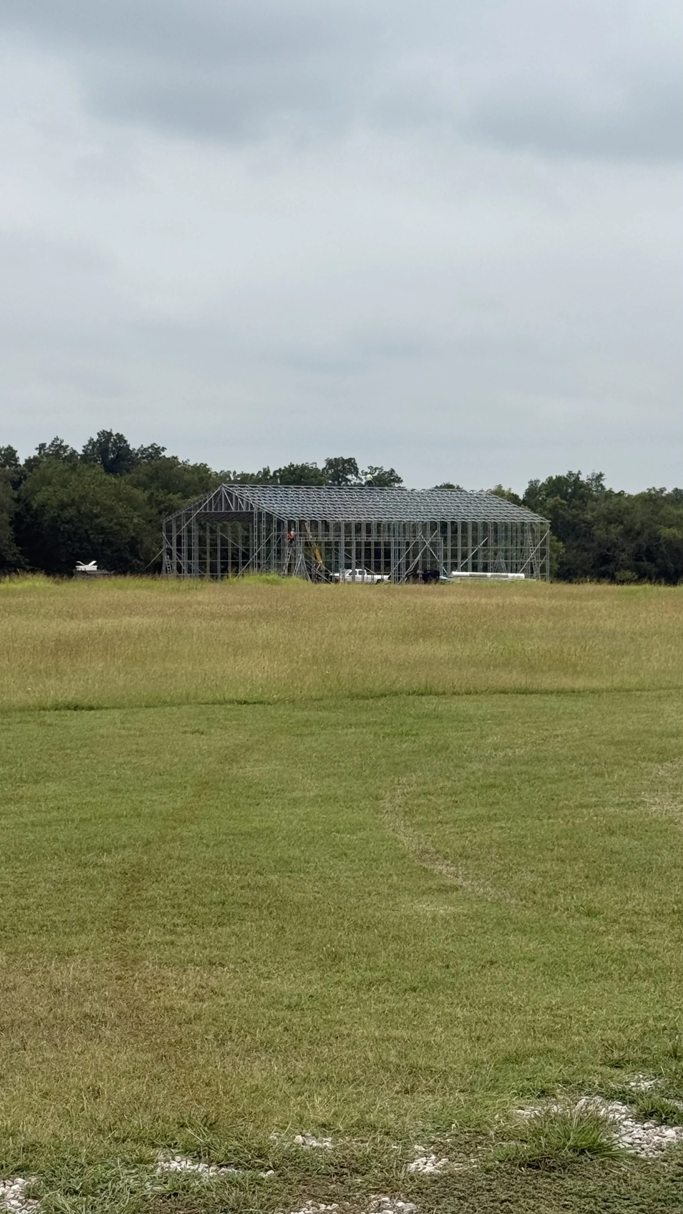 A grassy field with a large metal framework structure under construction in the distance, with trees and cloudy sky in the background.