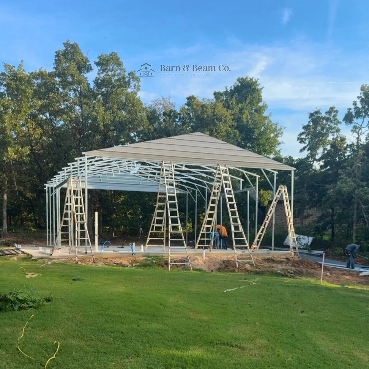 Construction of a metal-frame building with workers on site, surrounded by green grass and trees, under a clear blue sky.