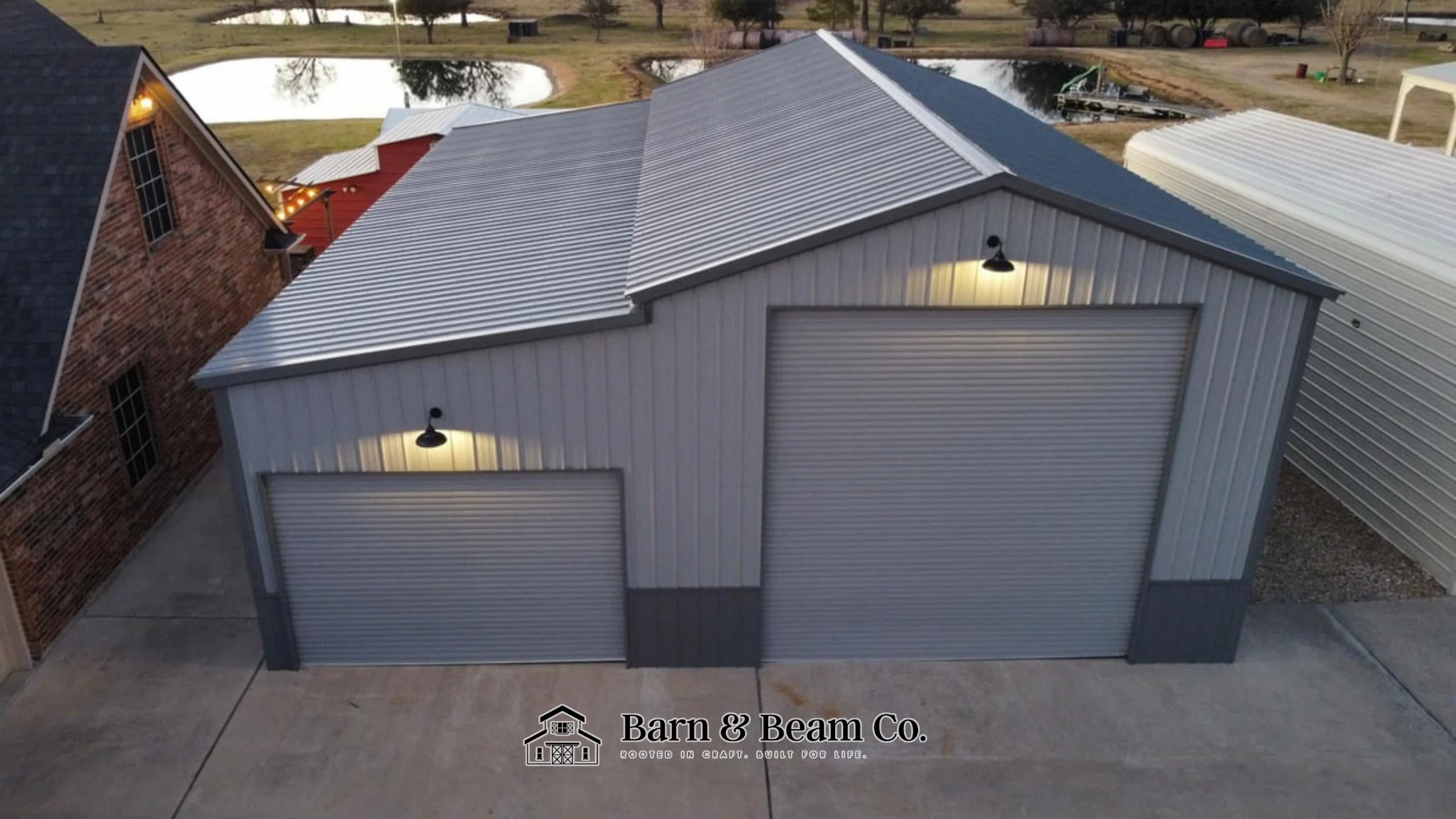 A metal storage building with two roll-up garage doors, exterior lights, and a metal roof, situated in a residential area with neighboring buildings and a body of water in the background.