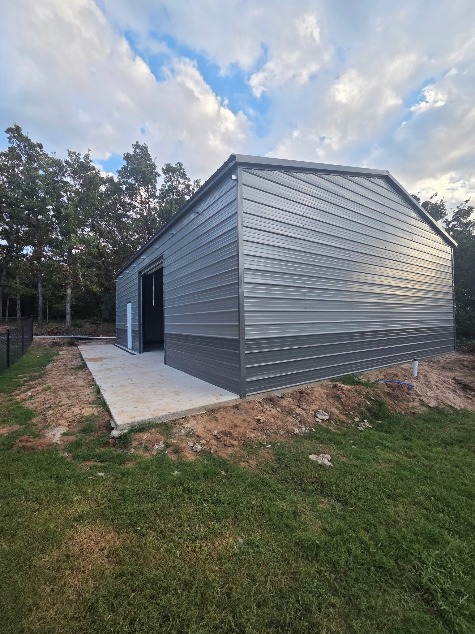 A metal storage shed on a concrete slab, surrounded by a grassy yard and trees under a partly cloudy sky.