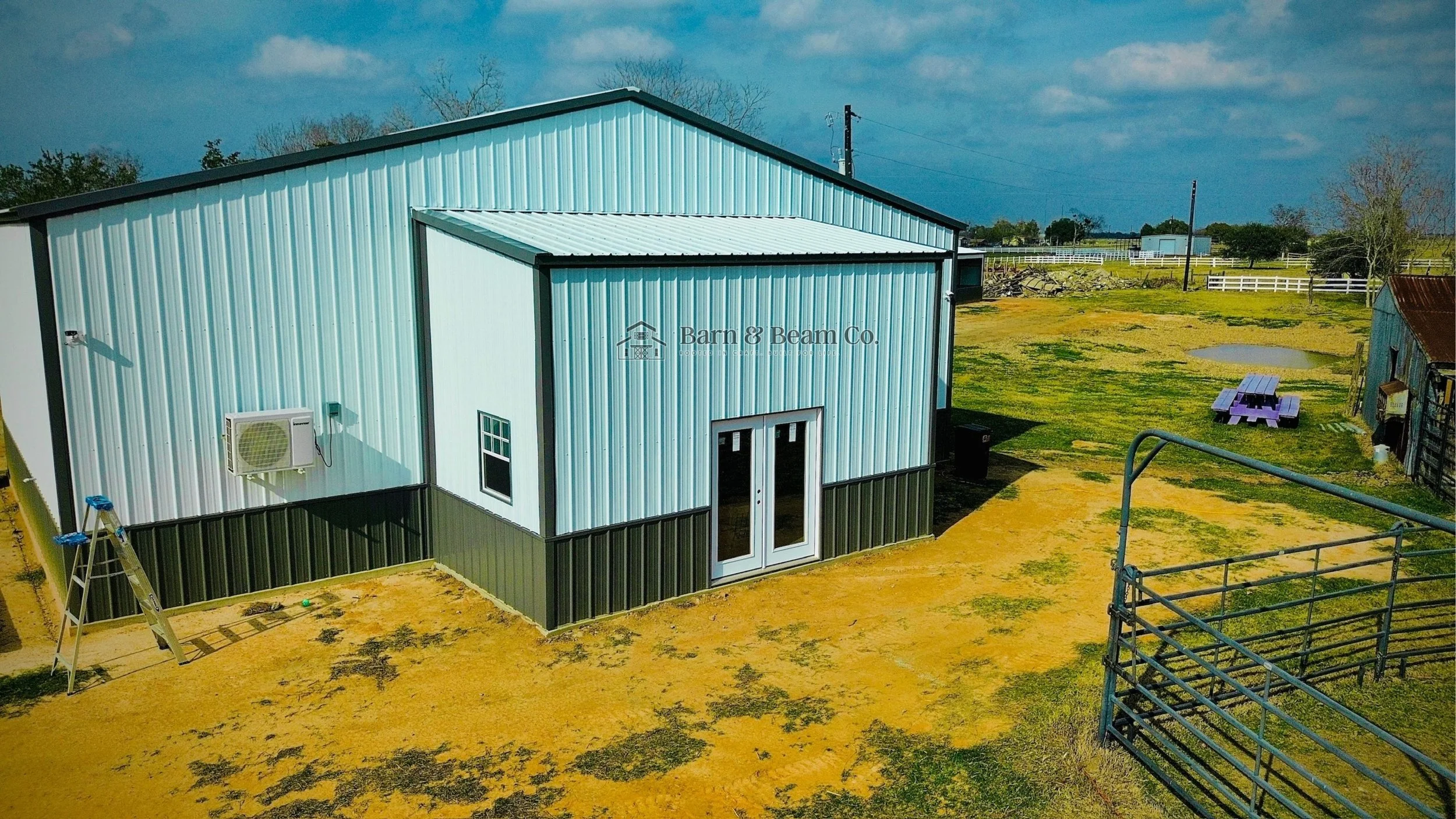 Photo of a large blue metal barn with double glass doors, a ladder leaning against the wall, and a small window. The barn is set on yellowish ground with some patches of grass, and there is a purple picnic table and a smaller structure with a rusted 