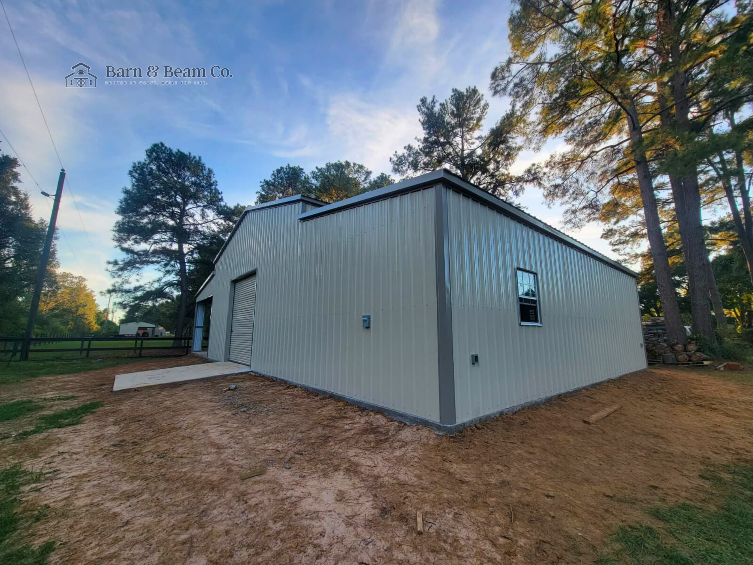 A large metal storage building with a roll-up door and a window on the side, situated on a patch of dirt with trees in the background.