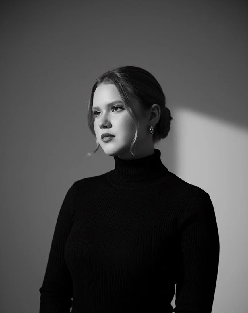 Retrato de uma mulher com cabelo preso, de cabelo longo e rosto delicado, usando um suéter preto de gola alta, em tom monocromático branco e preto, com expressão pensativa, iluminada por luz suave e sombra ao fundo.