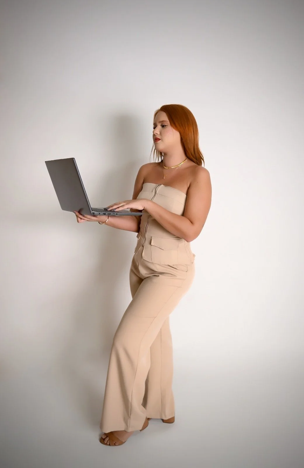 Mulher de cabelo ruivo e pele clara usando roupa bege, segurando um laptop, em pé contra um fundo branco.
