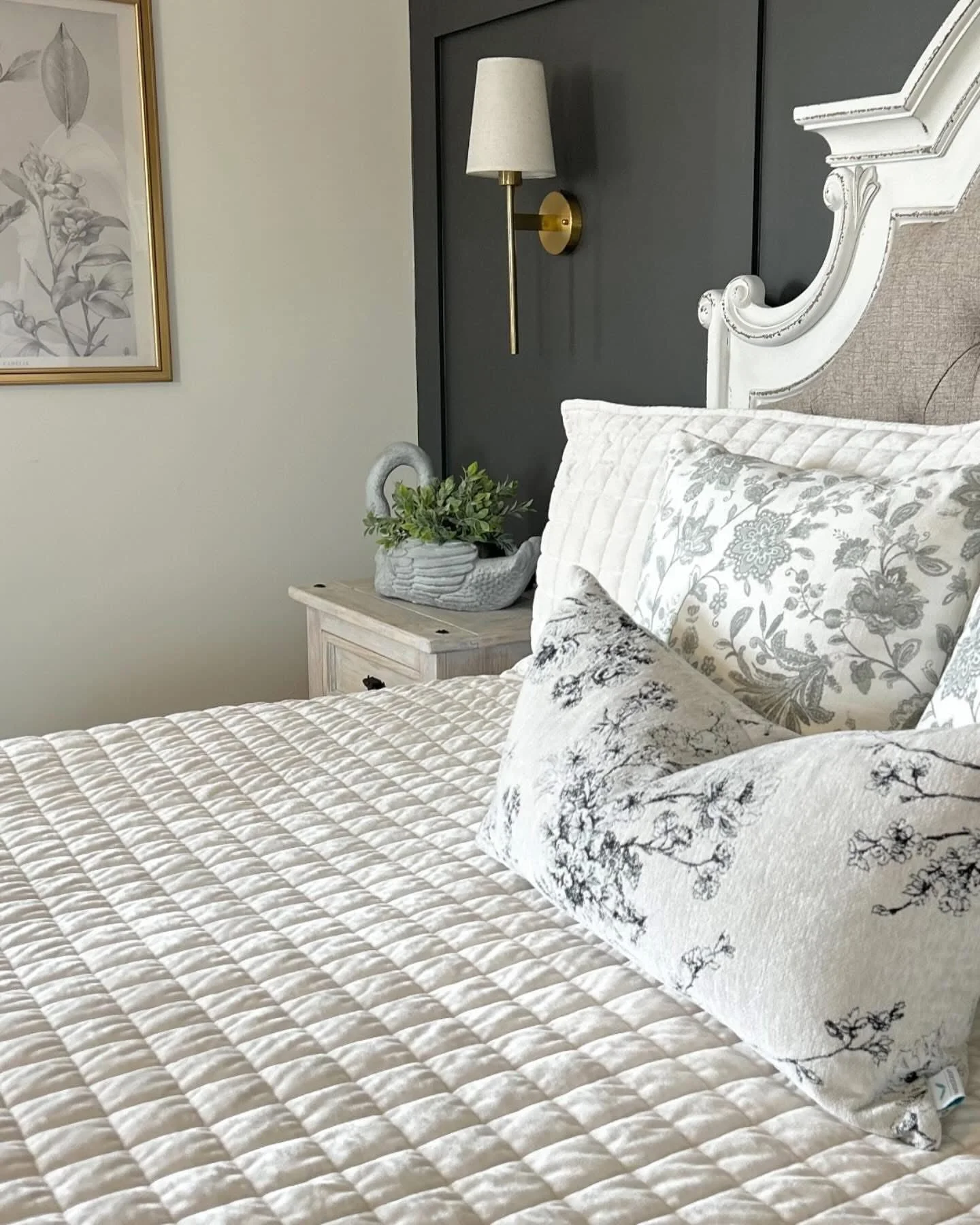 A neatly made bed with a white quilt, floral pillows, and a decorative pillow. Beside the bed is a nightstand with a plant in a swan-shaped pot. Camp Renovation and Design in Columbus, Ohio.