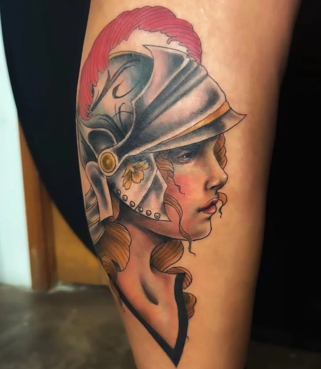 A detailed tattoo of a woman with wavy hair, wearing a classical helmet decorated with a red crest, and featuring realistic shading and color.