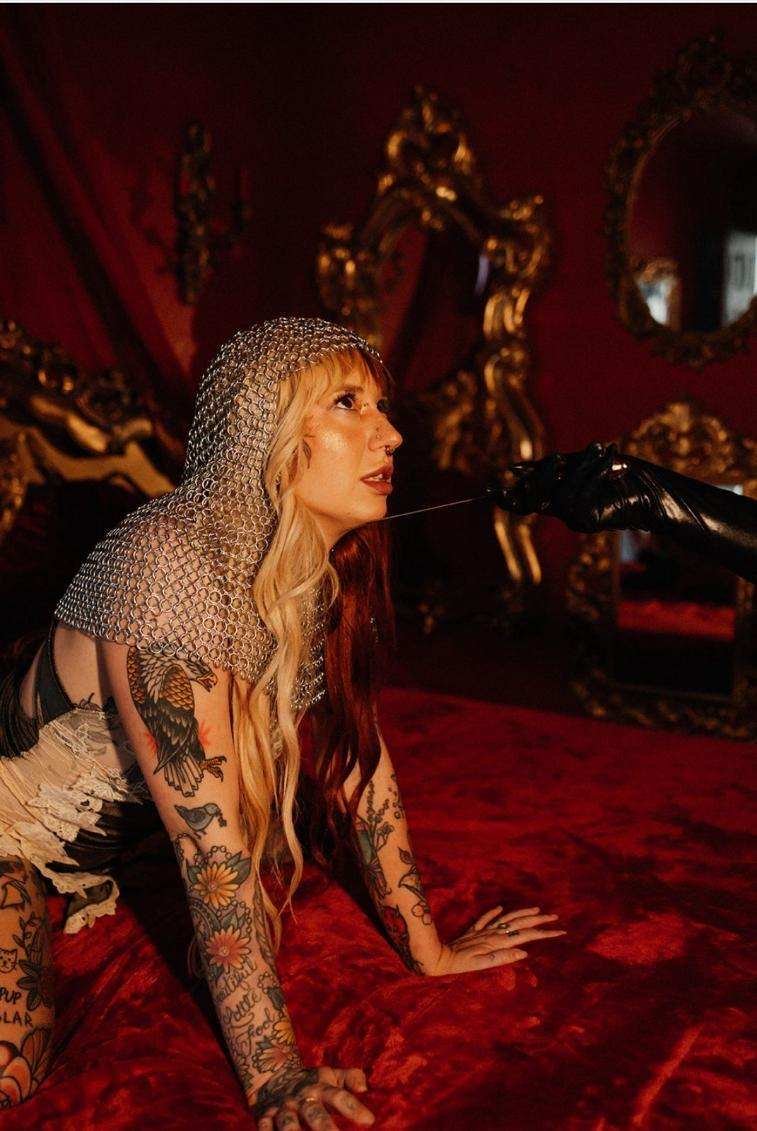 A woman with long tattoos on her arms and chest, wearing a chainmail hood, is on her hands and knees on a red velvet surface. A gloved hand is holding a thin string or thread near her mouth in a dimly lit room with ornate gold-framed mirrors and red walls.