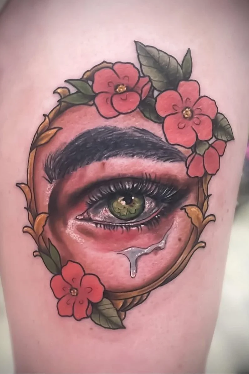 Tattoo of a realistic eye with green iris, tears, surrounded by red flowers and green leaves, outlined with a decorative brown frame.
