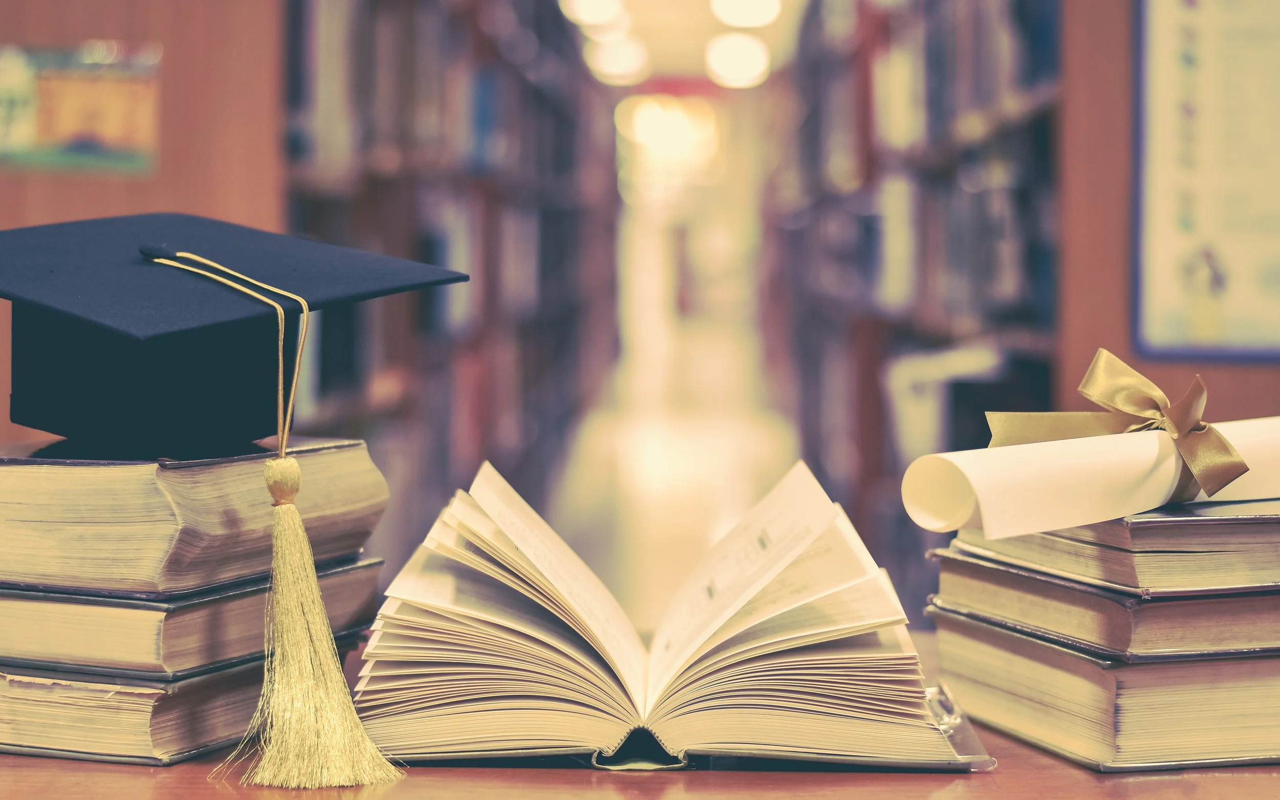 A graduation cap with a yellow tassel placed on an open book, surrounded by other books and diplomas with ribbons. The background shows a blurred library setting.