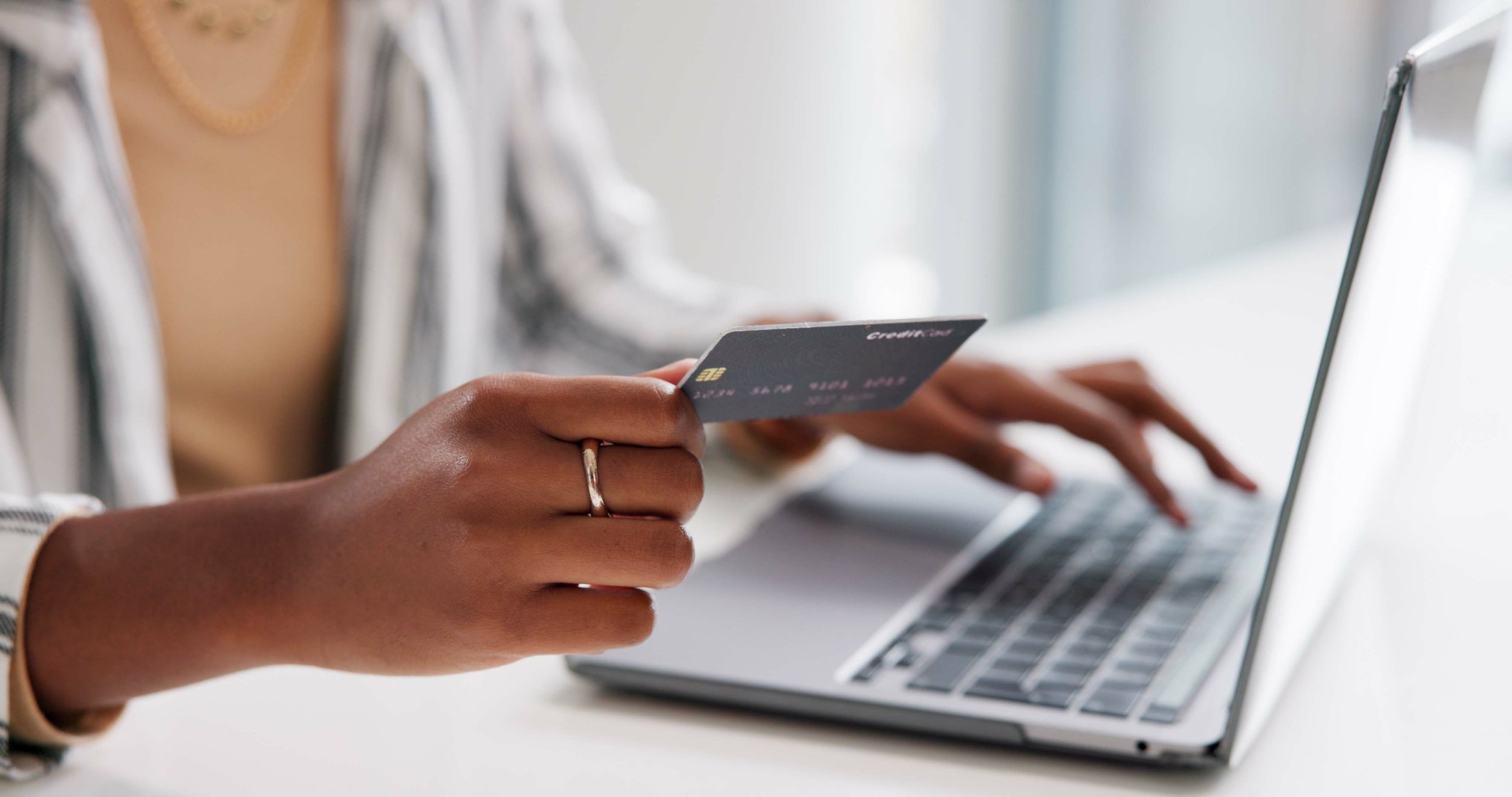 A person holding a credit card and typing on a laptop keyboard, sitting at a white desk.