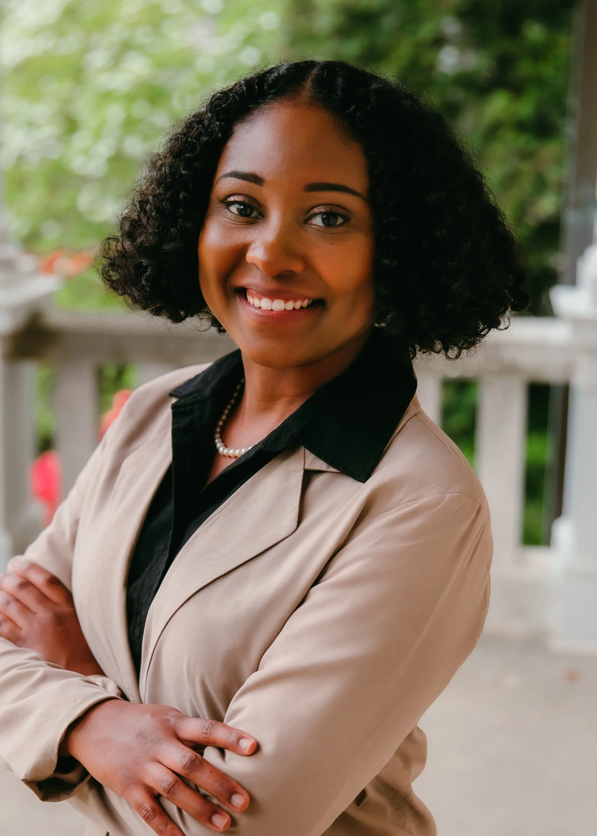Smiling woman with curly black hair, wearing a beige blazer, black shirt, pearl necklace, standing outdoors with greenery in background.