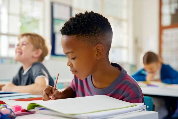 Young boy writing at a desk in a classroom with other children in the background.