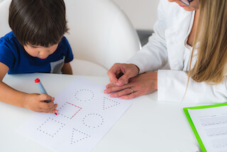 Child coloring on a sheet with dotted shapes, while an adult oversees, seated at a table.
