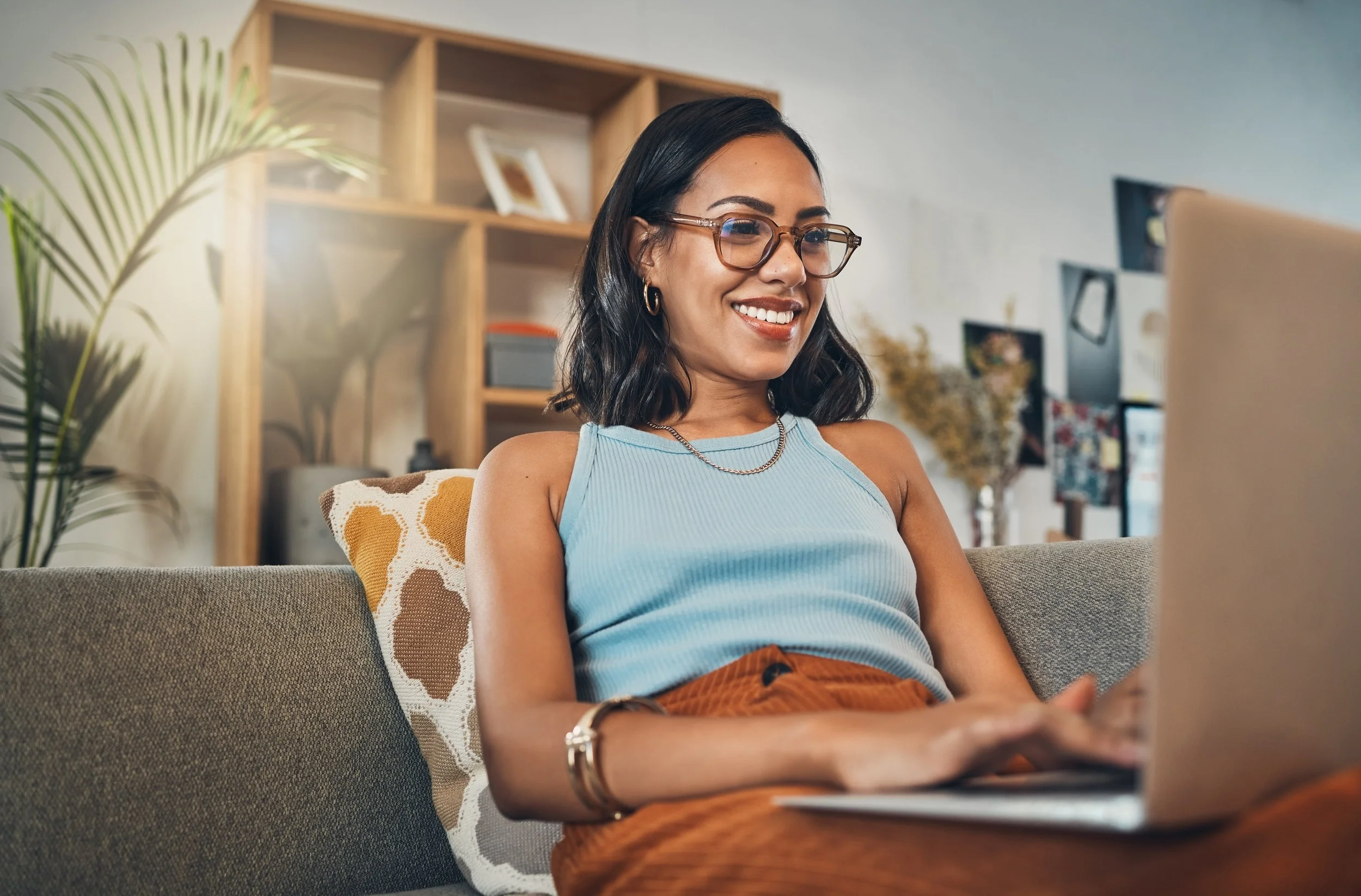 A woman with black hair, wearing glasses, a light blue sleeveless top, and gold jewelry, smiling while sitting on a gray sofa with a decorative cushion, using a laptop in a cozy, well-decorated living room with wooden shelves and various decorative items.