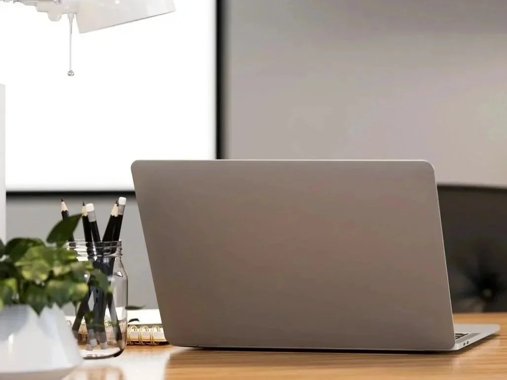 A closed silver laptop on a wooden desk surrounded by a potted plant, a glass jar with black pens, a small notepad with a gold spiral, and a white object. A computer monitor is visible in the background.