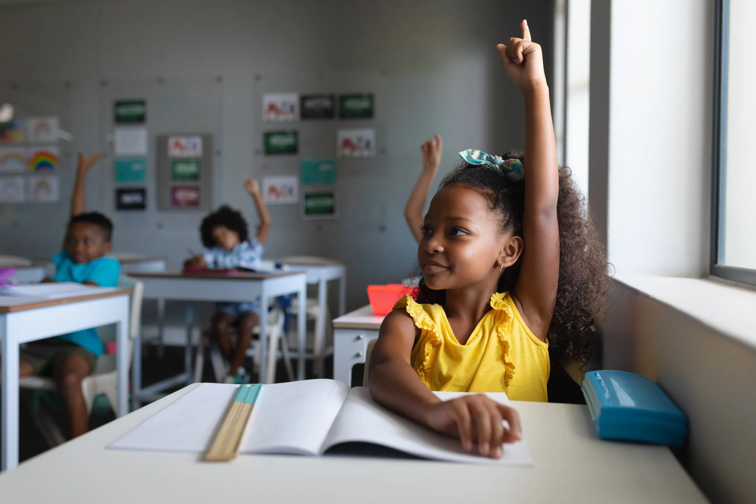 A young girl with curly hair wearing a yellow dress, sitting at her desk in a classroom, raising her hand with a smile. Other children are also seated at desks, raising their hands, with classroom decorations on the wall behind them.