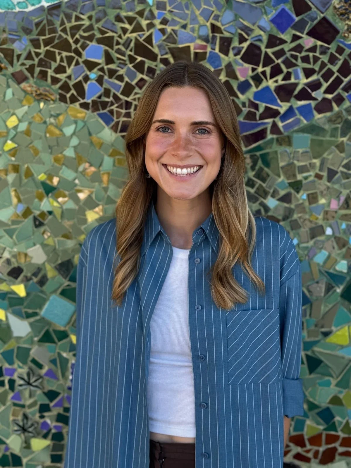 A young woman with long, wavy brown hair, blue eyes, and a bright smile, wearing a blue pinstripe shirt over a white top, standing in front of a colorful mosaic wall.