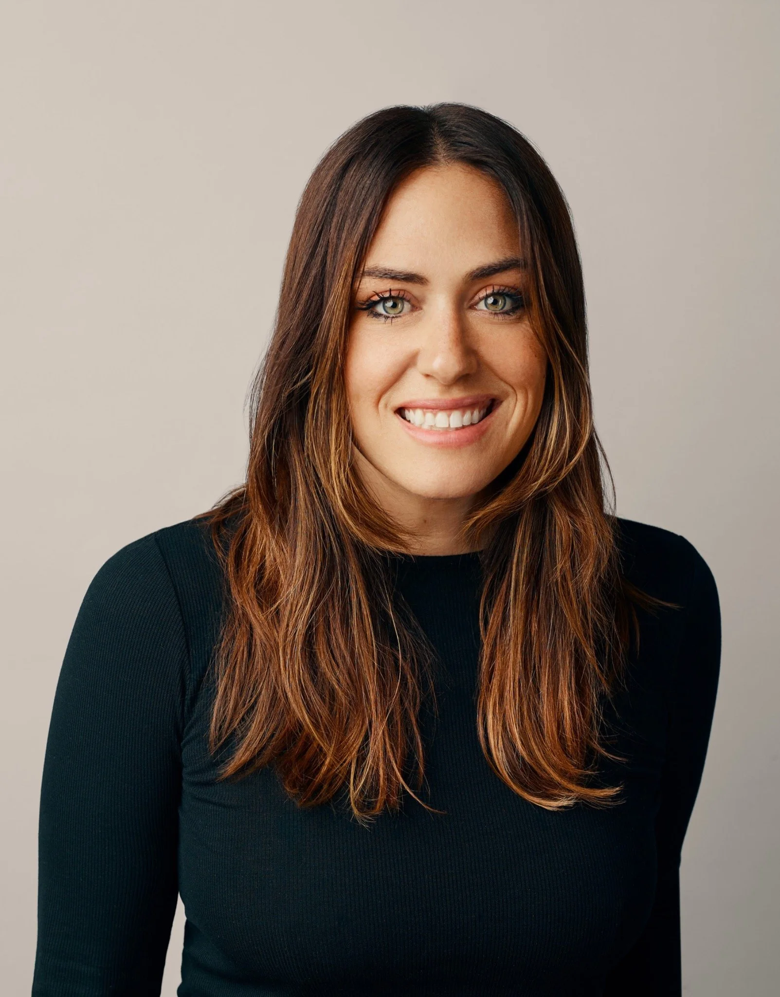 Portrait of a woman with long brown hair wearing a black top, smiling at the camera against a neutral background.