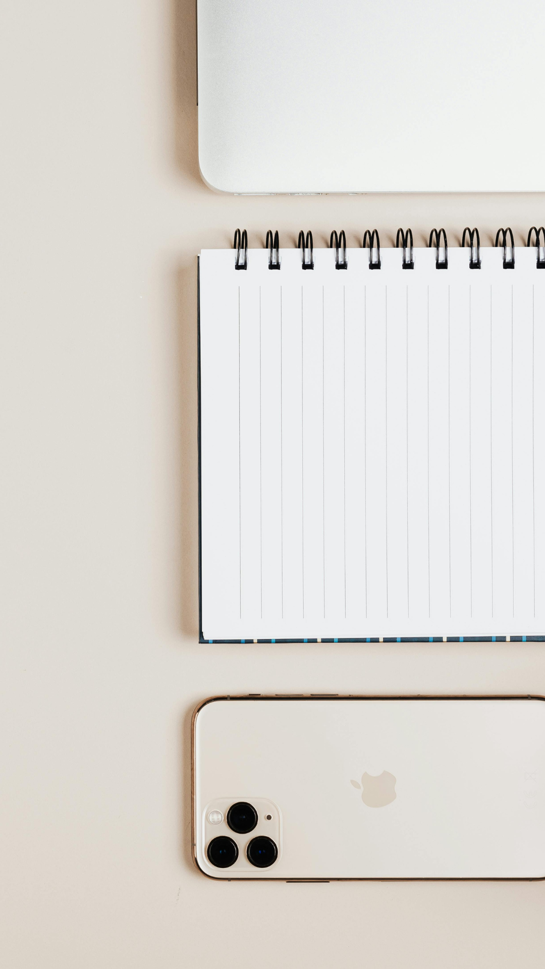 Top-down view of a closed silver laptop, a blank spiral notebook, and a white iPhone with three camera lenses on a beige surface.