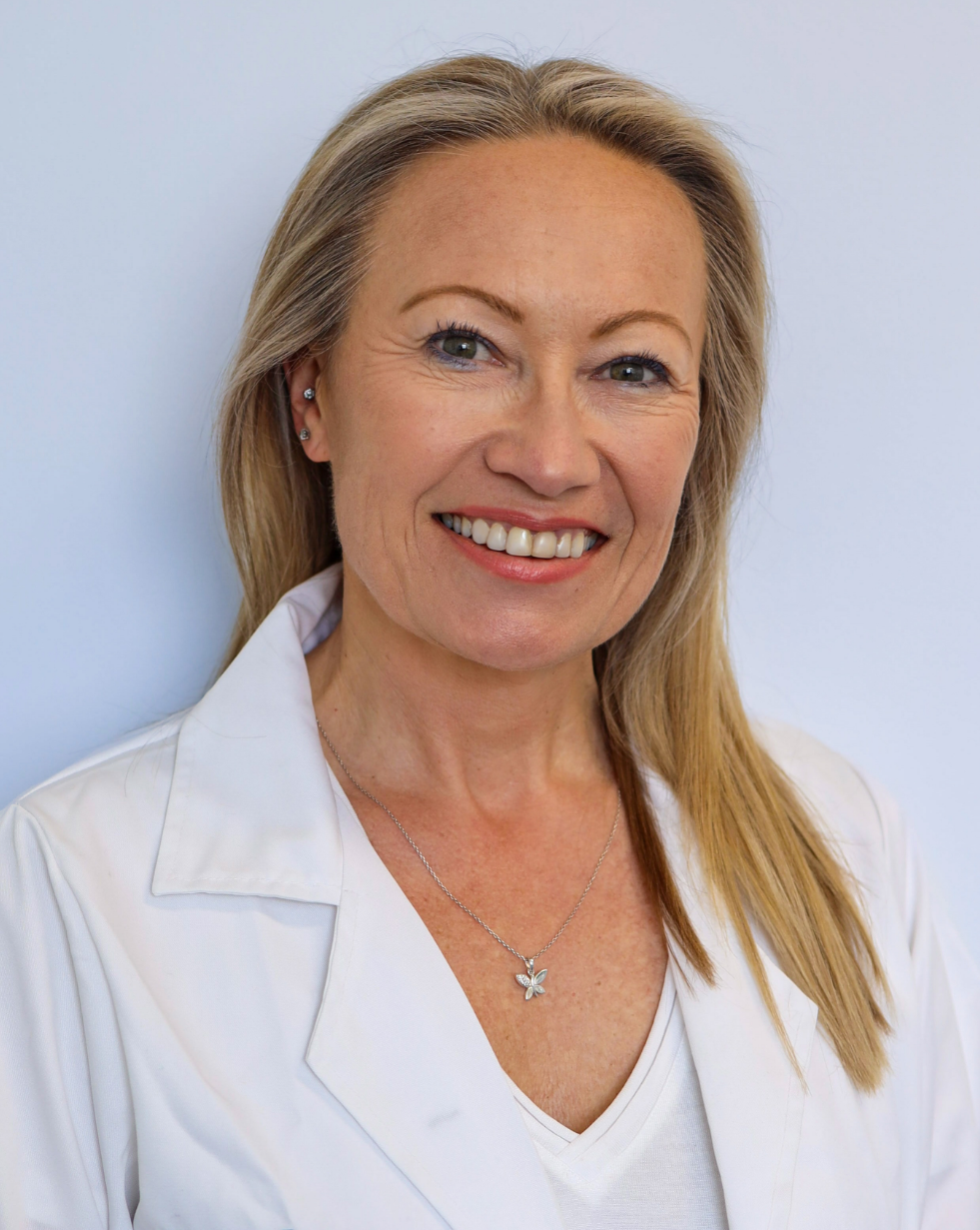 A smiling middle-aged woman with blonde hair, wearing a white shirt and a butterfly necklace, standing against a plain light background.