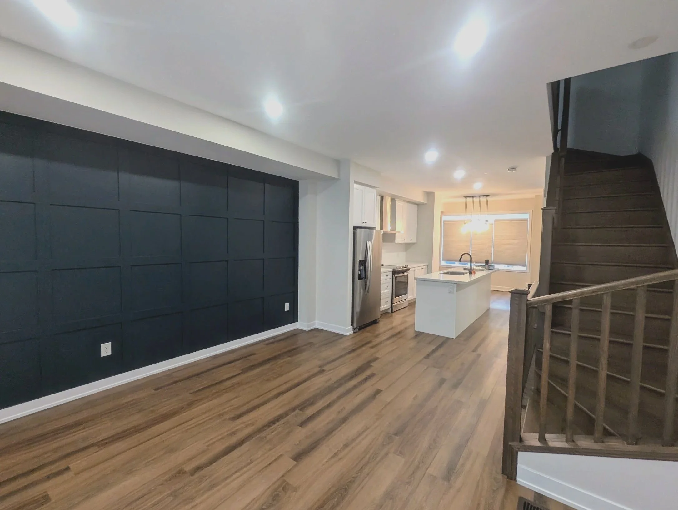 An open floor plan kitchen and living area with hardwood floors, a dark accent wall, white cabinetry, stainless steel refrigerator, and a staircase with wooden railing. beautiful trim,