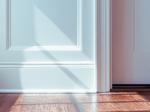 Close-up of a white door with shadows on the floor and wall, showing part of the door frame and hardwood flooring.