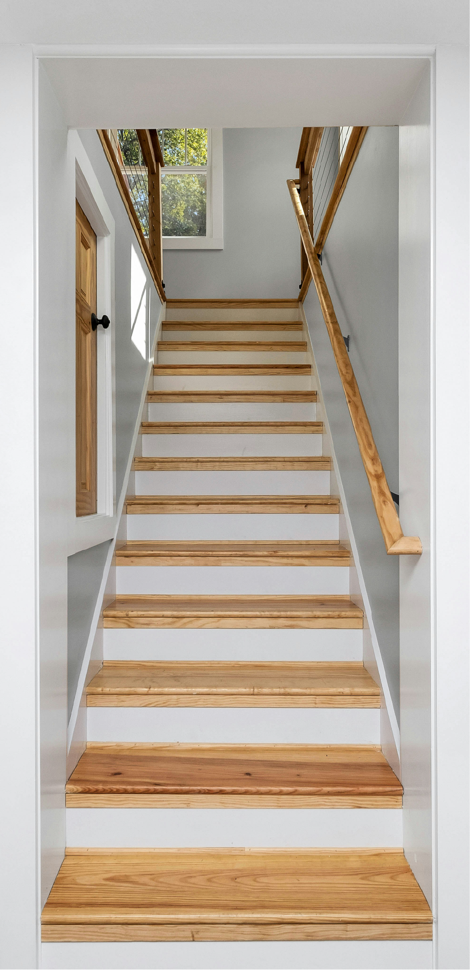 Wooden staircase with white risers, leading upward to a second floor with a wooden railing and an open window showing trees outside.