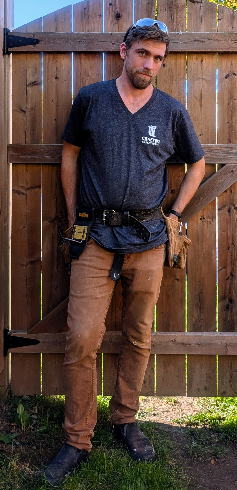 A man with a beard and glasses on his head standing outdoors in front of a wooden fence. He is wearing a dark gray t-shirt, brown work pants, and black work boots. He has a tool belt and a pouch on his side, and is leaning slightly with one hand in his pocket.