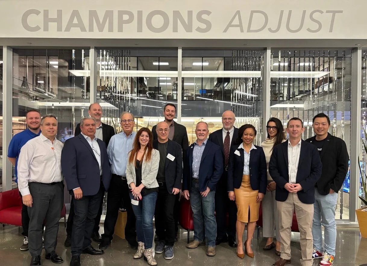 Group of 14 professionals posing for a photo in front of a glass-walled office or conference room at a business event or meeting.