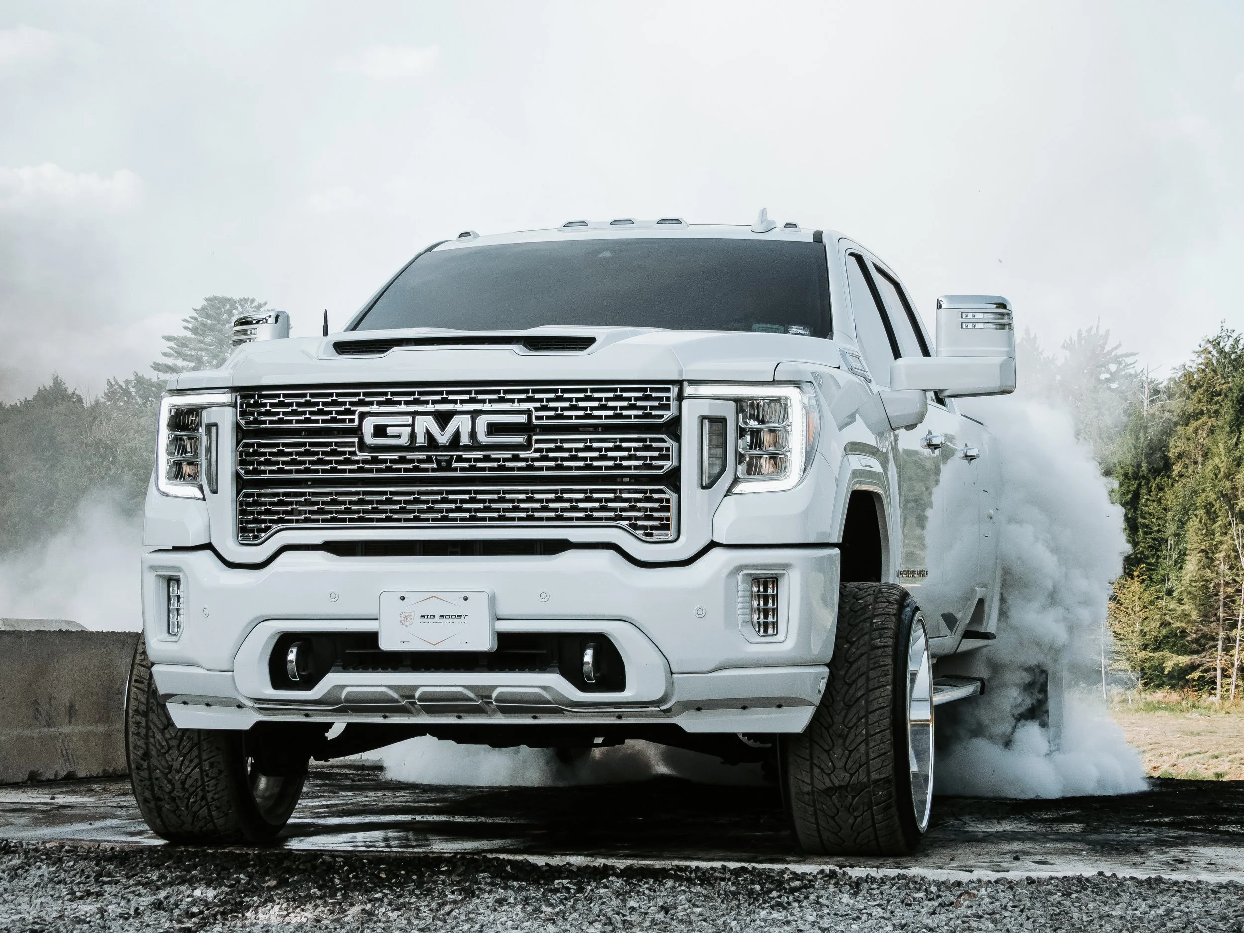 White GMC pickup truck performing a burnout, generating smoke from the rear tires on a rugged outdoor terrain with trees and a cloudy sky in the background.
