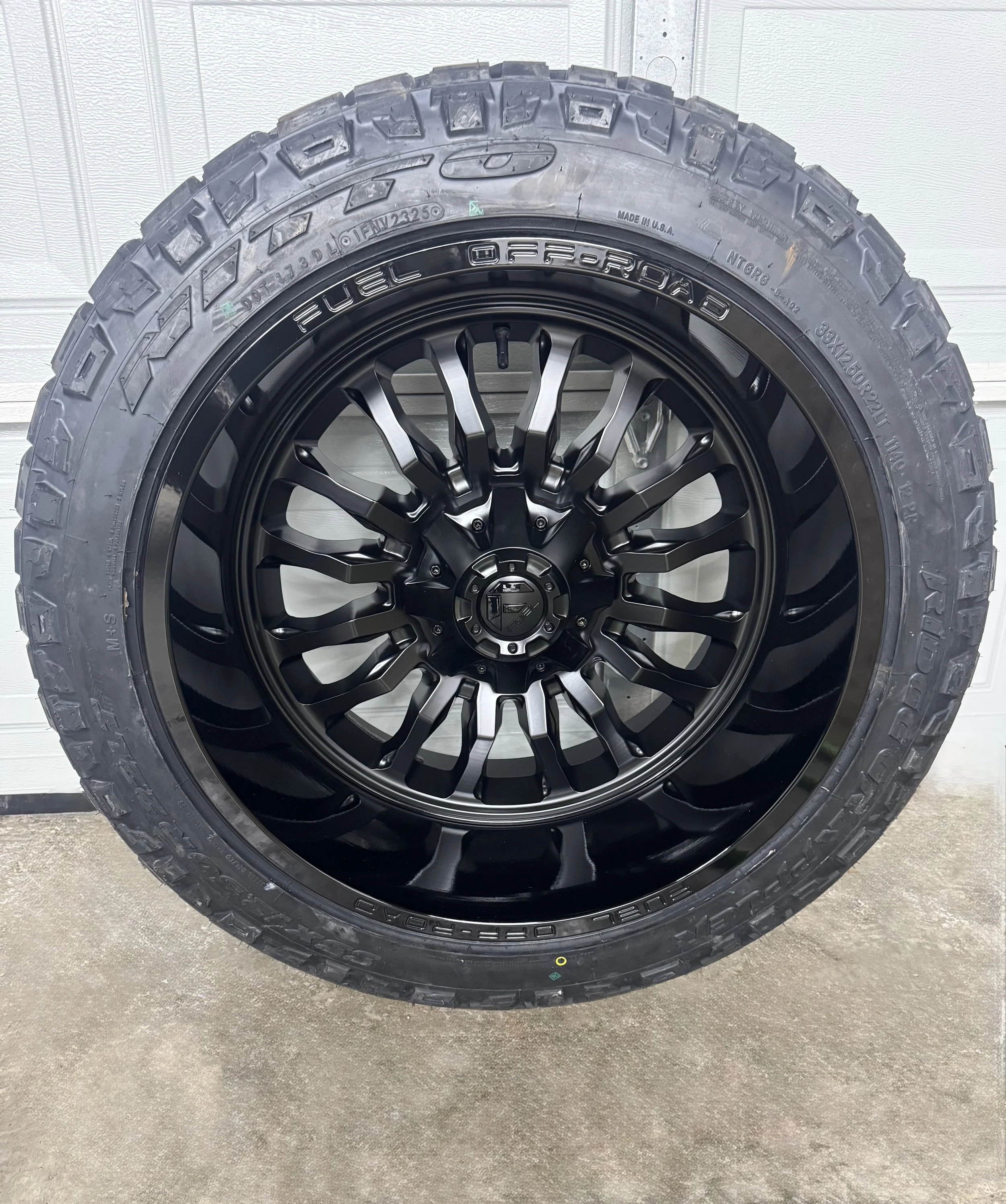 Black off-road tire with large tread and black rim with multiple spokes, mounted on a vehicle.