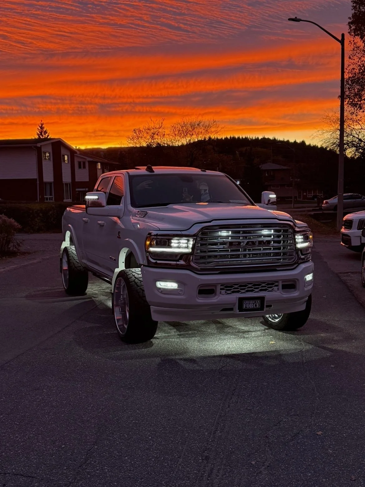 A lifted white RAM truck parked on an asphalt lot during sunset with a vibrant orange and pink sky, residential buildings, and trees in the background.