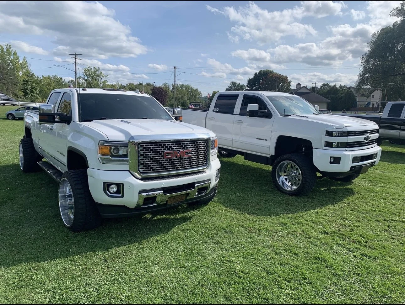 Two white GMC Sierra pickup trucks with lifted suspensions and custom rims parked on a grassy field, under a partly cloudy sky.