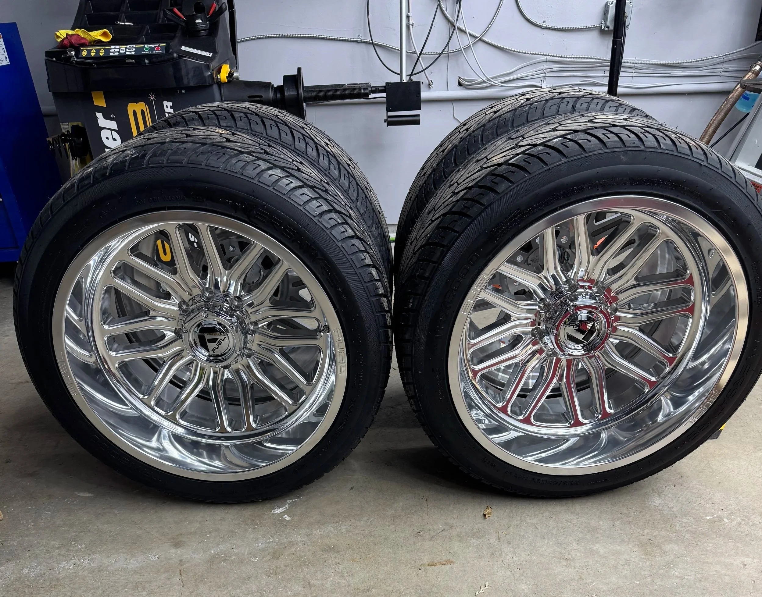 Two shiny chrome wheels with tires mounted, placed side by side on a concrete floor in a garage or workshop.