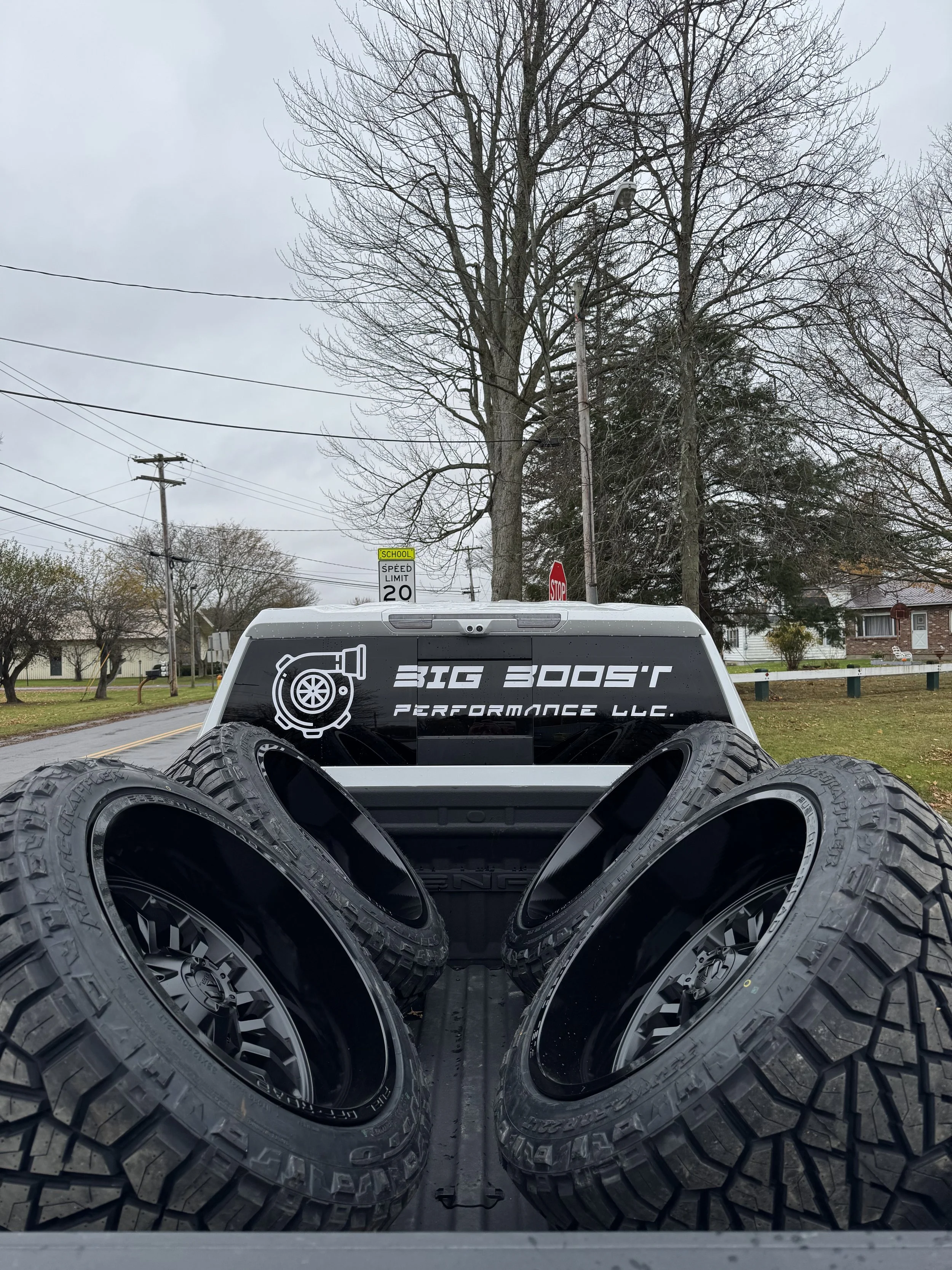Four large off-road tires in the truck bed of a pickup truck with a 'Big Boost Performance LLC' decal on the rear window. The scene is outdoors on a cloudy day with leafless trees and residential houses.