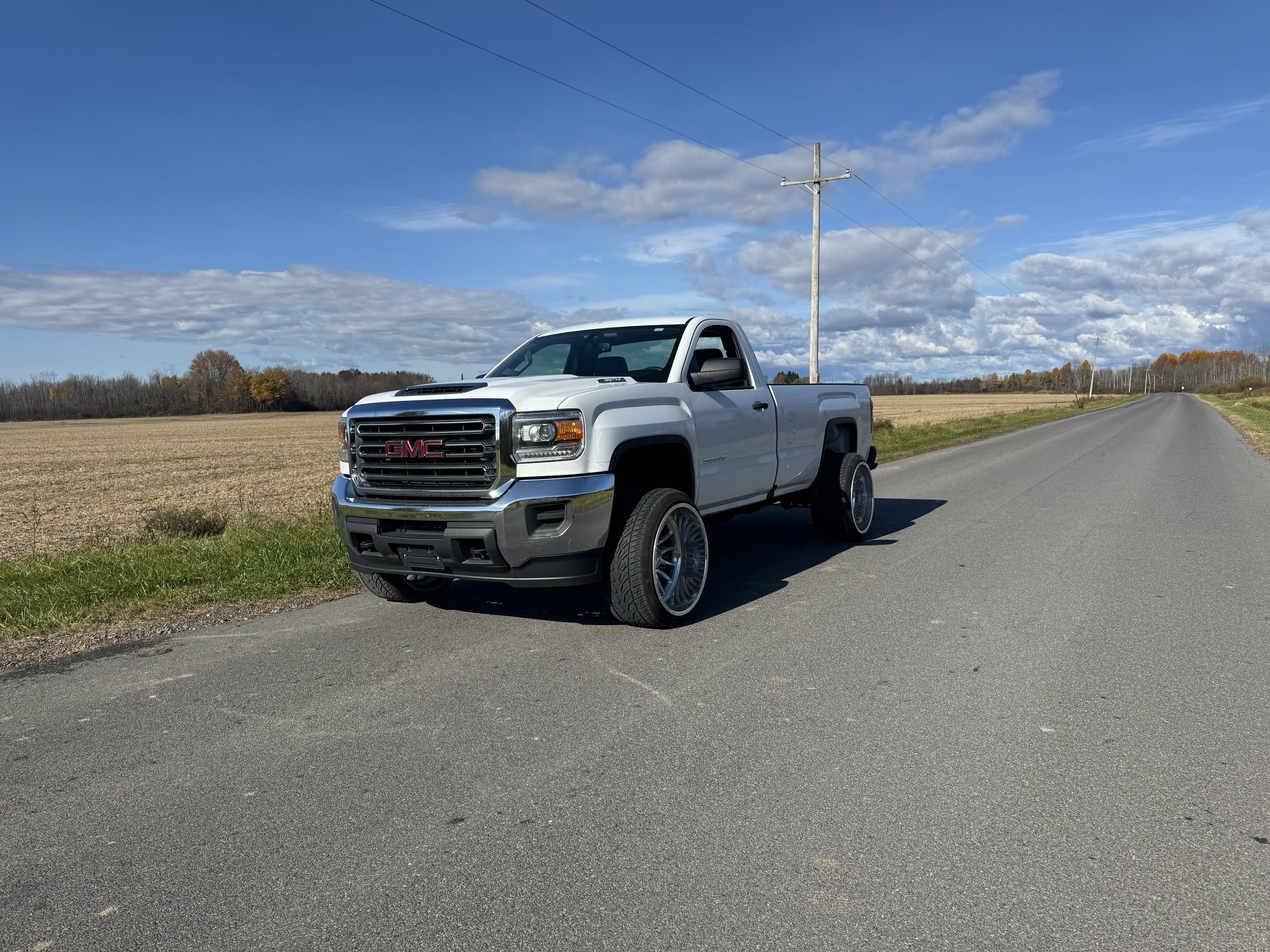 A white GMC pickup truck parked on the side of a rural road with open fields and a partly cloudy sky in the background.