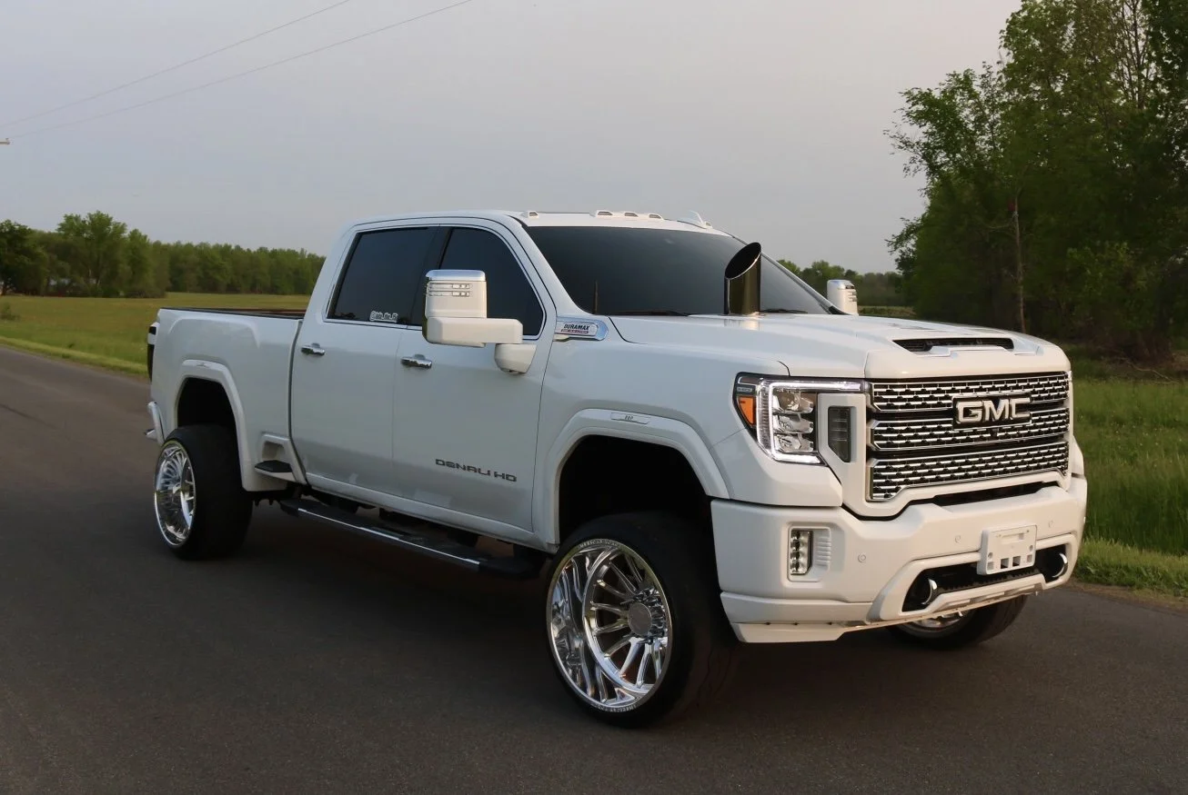 A white GMC pickup truck with large chrome wheels parked on the side of a rural road, with trees and grass in the background.