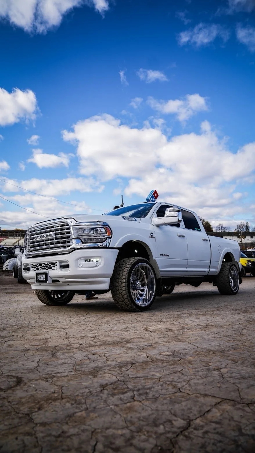 White lifted RAM pickup truck with custom wheels parked on cracked pavement under a partly cloudy sky during daytime.