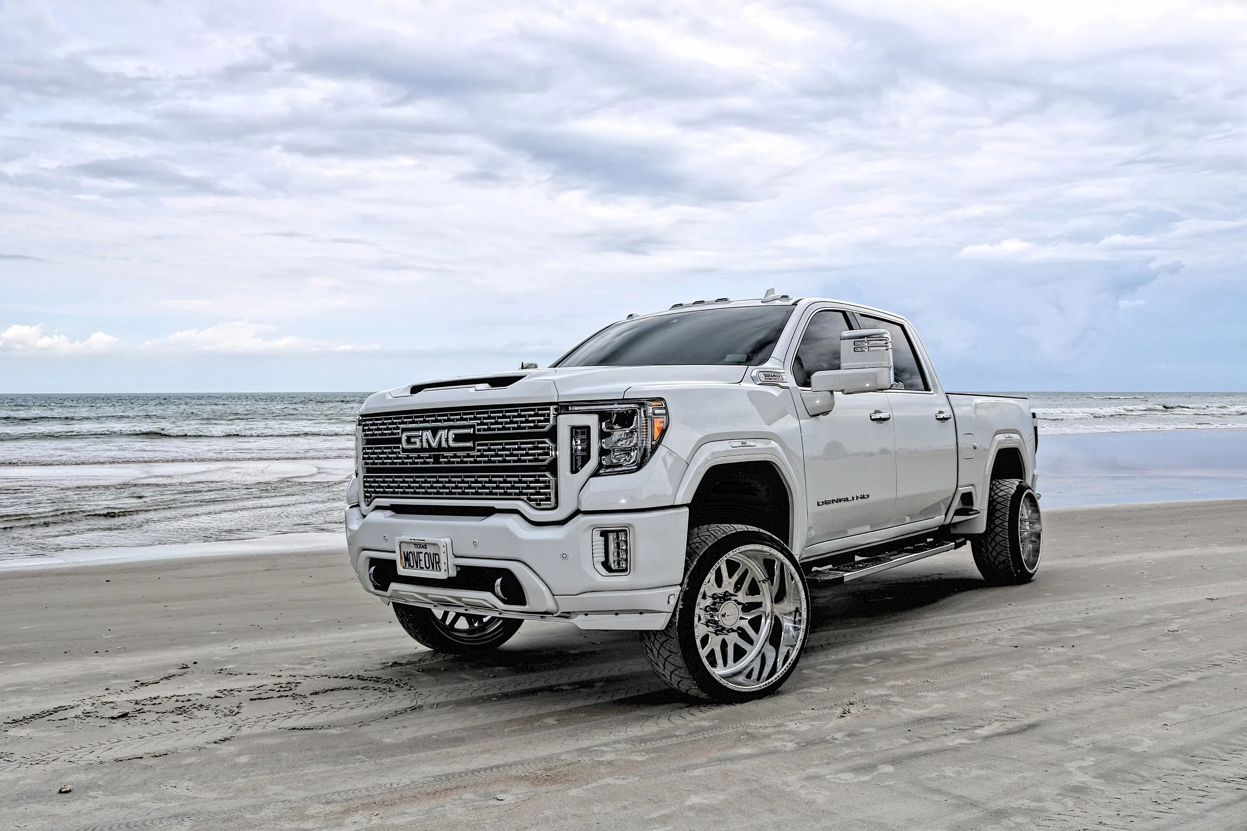 A white GMC Denali HD pickup truck with oversized wheels parked on a sandy beach near the ocean, under a cloudy sky.