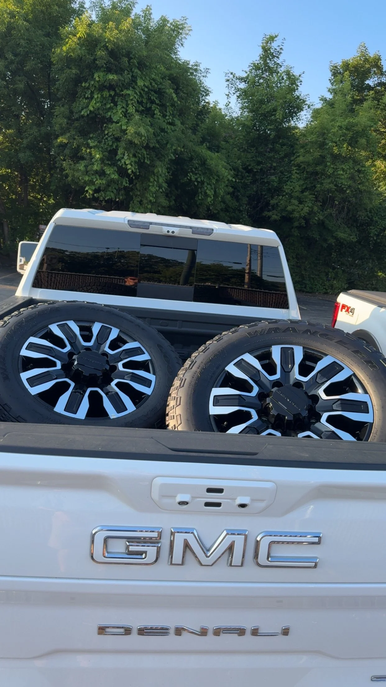 GMC Denali pickup truck with two large tires in the truck bed, parked outdoors with trees and blue sky in the background.