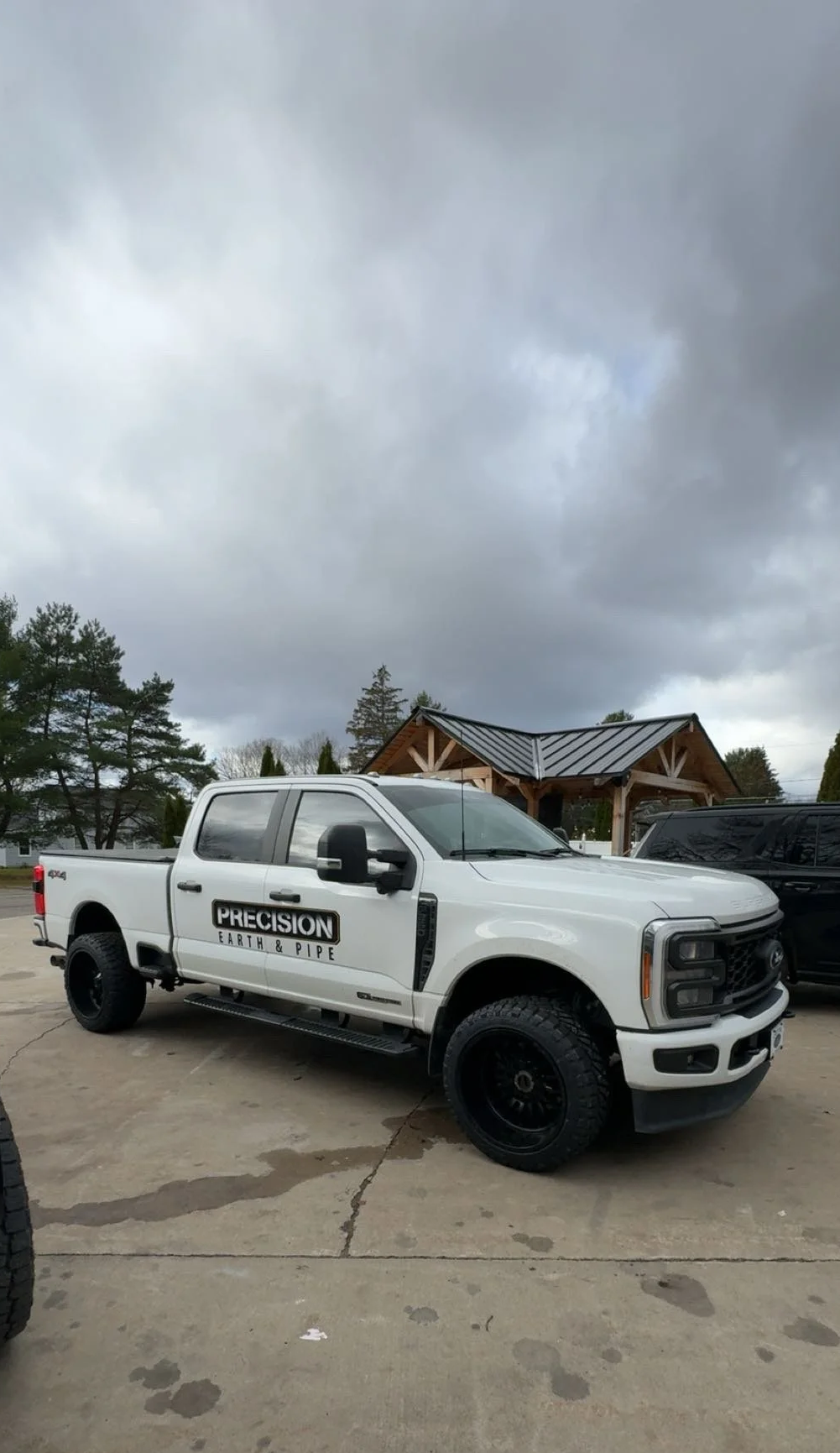 White pickup truck with black wheels and 'Precision Earth & Pipe' logo parked on a driveway under a cloudy sky, with a wooden structure in the background.
