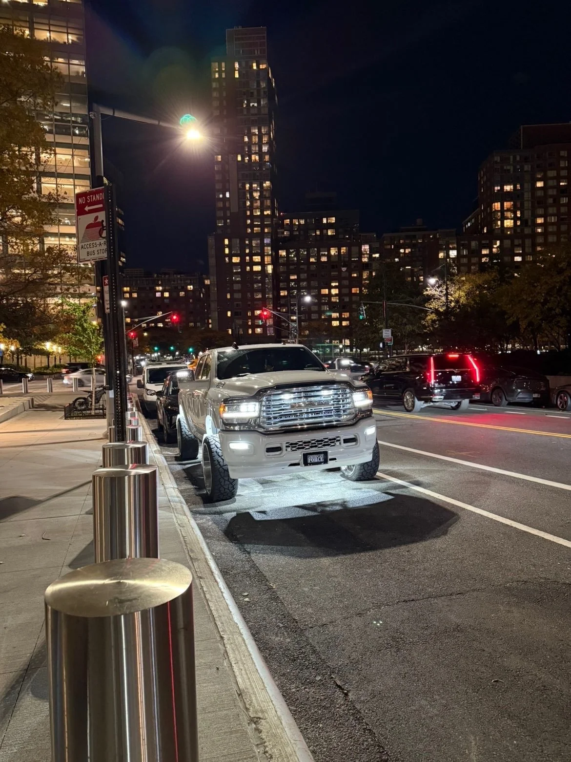 Nighttime city street with parked white pickup truck, illuminated by streetlights, with tall buildings and traffic in the background.