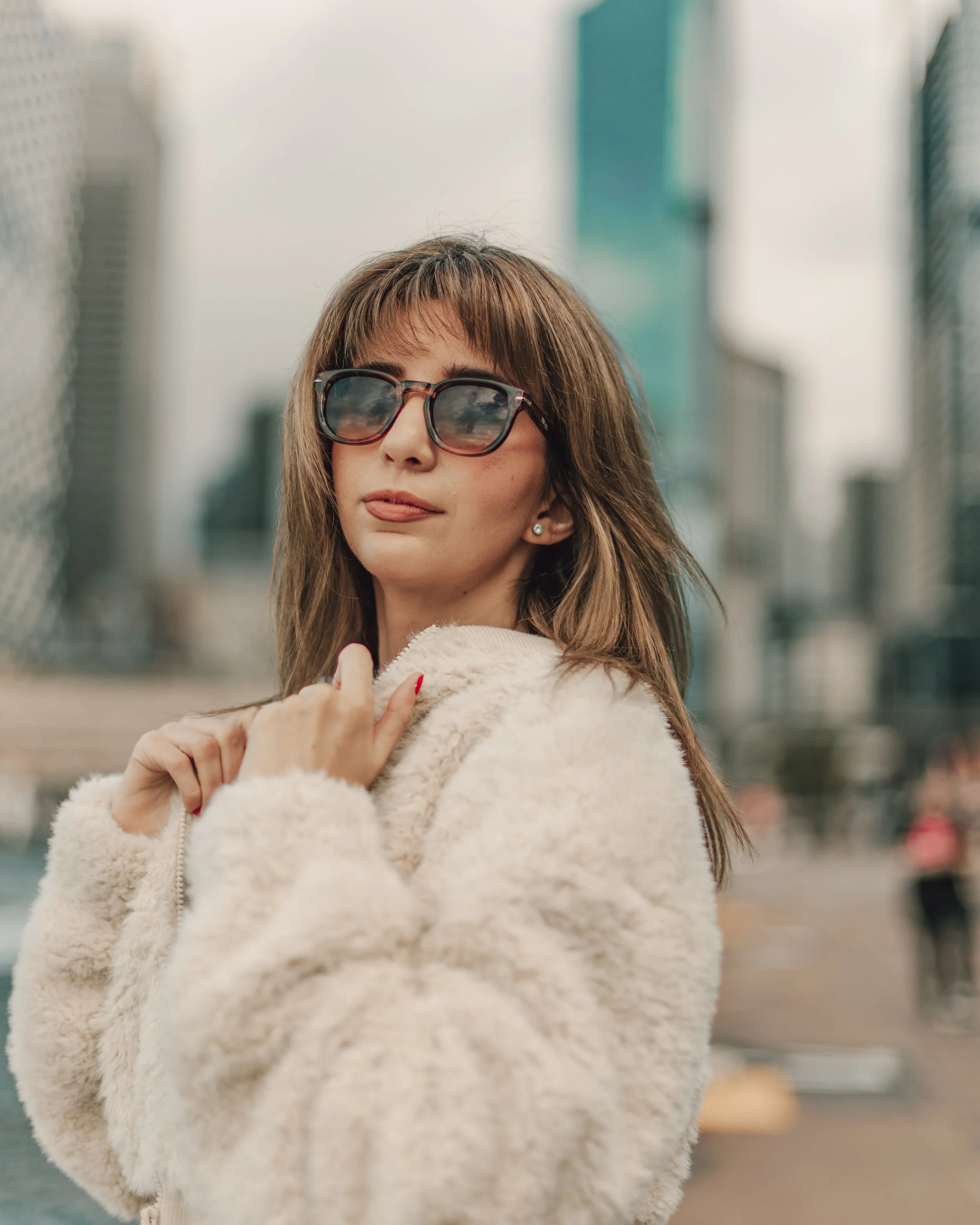 A woman with brown hair, wearing sunglasses and earrings, dressed in a cream-colored fuzzy jacket, standing outdoors in an urban environment with blurred buildings in the background.