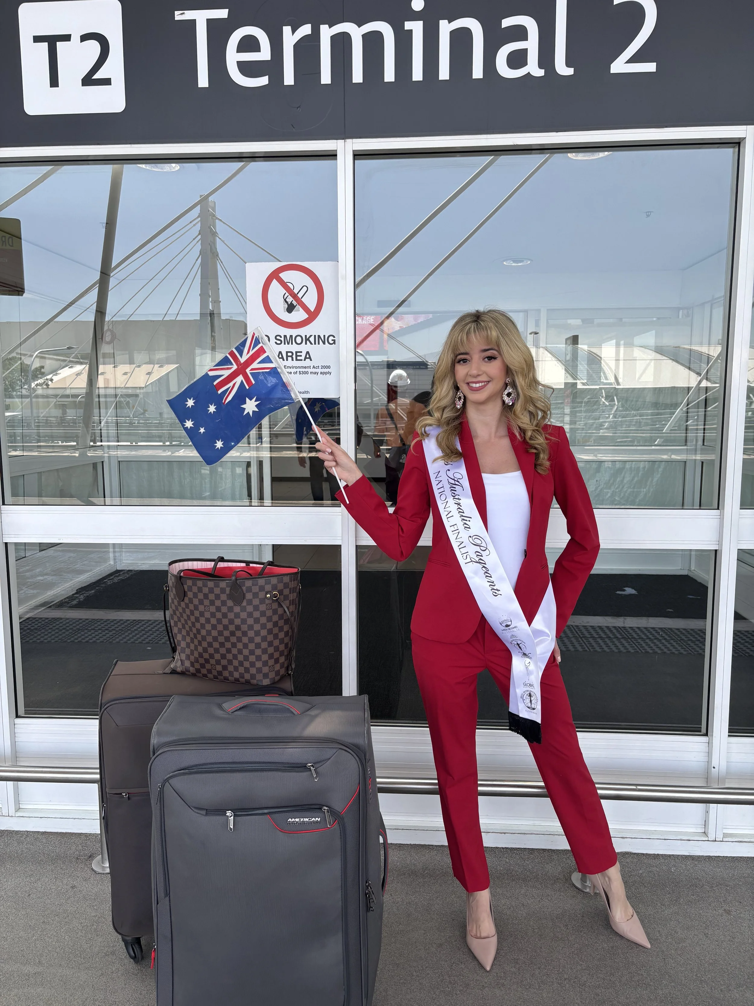 A woman in a red suit with a sash reading 'Miss Australia' is standing in front of an airport terminal holding an Australian flag. There are two suitcases and a handbag beside her, and a 'No Smoking' sign on the glass door behind her.